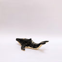 A small whale-shaped ceramic sculpture with a matte black finish and sgraffito beige details, including a beige underside and a gold circular accent on its side, set against a plain light gray background.