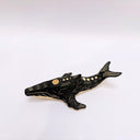 A small whale-shaped ceramic sculpture with a matte black finish and sgraffito beige details, including a beige underside and a gold circular accent on its side, set against a plain light gray background.