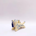 Cream colored ceramic sculpture of a wolf with a small cream colored frog on its back. Both have blue colored floral design motifs on the sides of their bodies. The wolf holds a dark blue tassel in its mouth.