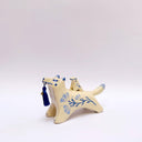 Cream colored ceramic sculpture of a wolf with a small cream colored frog on its back. Both have blue colored floral design motifs on the sides of their bodies. The wolf holds a dark blue tassel in its mouth.