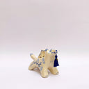 Cream colored ceramic sculpture of a wolf with a small cream colored frog on its back. Both have blue colored floral design motifs on the sides of their bodies. The wolf holds a dark blue tassel in its mouth.