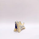 Cream colored ceramic sculpture of a wolf with a small cream colored frog on its back. Both have blue colored floral design motifs on the sides of their bodies. The wolf holds a dark blue tassel in its mouth.