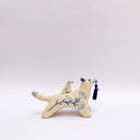 Cream colored ceramic sculpture of a wolf with a small cream colored frog on its back. Both have blue colored floral design motifs on the sides of their bodies. The wolf holds a dark blue tassel in its mouth.