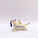 Cream colored ceramic sculpture of a wolf with a small cream colored frog on its back. Both have blue colored floral design motifs on the sides of their bodies. The wolf holds a dark blue tassel in its mouth.