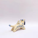 Cream colored ceramic sculpture of a wolf with a small cream colored frog on its back. Both have blue colored floral design motifs on the sides of their bodies. The wolf holds a dark blue tassel in its mouth.