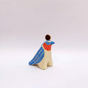 A ceramic bird sculpture with a natural cream colored clay body, blue wings and face detailing, red neck feathers, and a gold circular topper on its head, photographed on a clean light gray studio background.