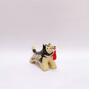 Black and cream colored ceramic sculpture of a husky dog, with a dog pack illustration on the side of its body. It holds a red tassel in its mouth with a small bronze bell.