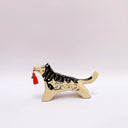Black and cream colored ceramic sculpture of a husky dog, with a dog pack illustration on the side of its body. It holds a red tassel in its mouth with a small bronze bell.