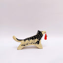 Black and cream colored ceramic sculpture of a husky dog, with a dog pack illustration on the side of its body. It holds a red tassel in its mouth with a small bronze bell.