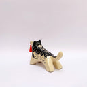 Black and cream colored ceramic sculpture of a husky dog, with a dog pack illustration on the side of its body. It holds a red tassel in its mouth with a small bronze bell.