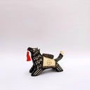 Ceramic sculpture of a black wolf with sgraffito cream colored fur lines, holding a red tassel in its mouth. On its back rests a cream colored blanket with a doll charm laying atop it.