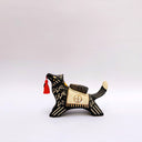 Ceramic sculpture of a black wolf with sgraffito cream colored fur lines, holding a red tassel in its mouth. On its back rests a cream colored blanket with a doll charm laying atop it.