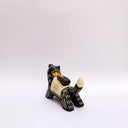 Ceramic sculpture of a black wolf with sgraffito cream colored fur lines, holding a red tassel in its mouth. On its back rests a cream colored blanket with a doll charm laying atop it.