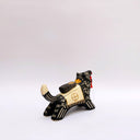 Ceramic sculpture of a black wolf with sgraffito cream colored fur lines, holding a red tassel in its mouth. On its back rests a cream colored blanket with a doll charm laying atop it.