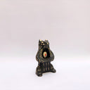 Black and cream colored ceramic sculpture of a bear, sitting on its hind legs with its arms cradled together. It has a recession in its chest where an oval shaped pennant with a red heart hangs. 