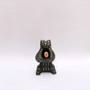 Black and cream colored ceramic sculpture of a bear, sitting on its hind legs with its arms cradled together. It has a recession in its chest where an oval shaped pennant with a red heart hangs. 