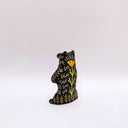 Black and cream colored ceramic sculpture of a bear, sitting on its hind legs with its arms cradled together. It has a recession in its chest where an oval shaped pennant with a red heart hangs. 