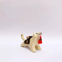 Ceramic sculpture of a cream colored wolf, with a red tassel hanging from its mouth and a black and gold blanket on its back. Atop the blanket is a wrapped doll charm.