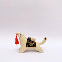 Ceramic sculpture of a cream colored wolf, with a red tassel hanging from its mouth and a black and gold blanket on its back. Atop the blanket is a wrapped doll charm.