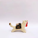 Ceramic sculpture of a cream colored wolf, with a red tassel hanging from its mouth and a black and gold blanket on its back. Atop the blanket is a wrapped doll charm.