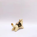Ceramic sculpture of a cream colored wolf, with a red tassel hanging from its mouth and a black and gold blanket on its back. Atop the blanket is a wrapped doll charm.