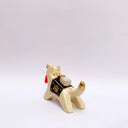Ceramic sculpture of a cream colored wolf, with a red tassel hanging from its mouth and a black and gold blanket on its back. Atop the blanket is a wrapped doll charm.