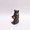 Black and cream colored ceramic sculpture of a bear, sitting on its hind legs with its arms cradled together. It has a recession in its chest where a gold crescent moon hangs. 
