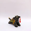 A black ceramic figurine of a wolf carrying a red tassel charm in its mouth, decorated with yellow and orange floral patterns and a small wrapped doll charm on its back, set against a plain light gray background.