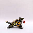 A black ceramic figurine of a wolf carrying a red tassel charm in its mouth, decorated with yellow and orange floral patterns and a small wrapped doll charm on its back, set against a plain light gray background.