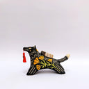 A black ceramic figurine of a wolf carrying a red tassel charm in its mouth, decorated with yellow and orange floral patterns and a small wrapped doll charm on its back, set against a plain light gray background.