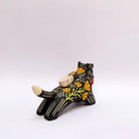 A black ceramic figurine of a wolf carrying a red tassel charm in its mouth, decorated with yellow and orange floral patterns and a small wrapped doll charm on its back, set against a plain light gray background.
