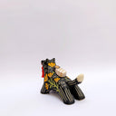 A black ceramic figurine of a wolf carrying a red tassel charm in its mouth, decorated with yellow and orange floral patterns and a small wrapped doll charm on its back, set against a plain light gray background.