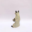 Ceramic sculpture of a cream and gray fox with black accent colorings, sitting up on its hind legs with its arms together in a cradling pose. Resting in its arms is a small wrapped doll.