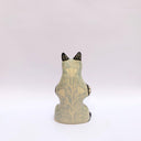 Ceramic sculpture of a cream and gray fox with black accent colorings, sitting up on its hind legs with its arms together in a cradling pose. Resting in its arms is a small wrapped doll.