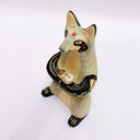 Ceramic sculpture of a cream and gray fox with black accent colorings, sitting up on its hind legs with its arms together in a cradling pose. Resting in its arms is a small wrapped doll.