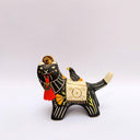 Ceramic sculpture of a black wolf with sgraffito line art for its fur and patterning. It hold a red tassel and charm in its mouth and has a blanket on its back. Sitting on the blanket is a small bird.