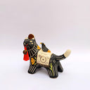 Ceramic sculpture of a black wolf with sgraffito line art for its fur and patterning. It hold a red tassel and charm in its mouth and has a blanket on its back. Sitting on the blanket is a small bird.