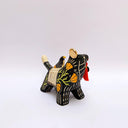 Ceramic sculpture of a black wolf with sgraffito line art for its fur and patterning. It hold a red tassel and charm in its mouth and has a blanket on its back. Sitting on the blanket is a small bird.