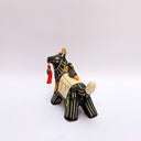 Ceramic sculpture of a black wolf with sgraffito line art for its fur and patterning. It hold a red tassel and charm in its mouth and has a blanket on its back. Sitting on the blanket is a small bird.
