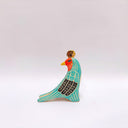 A stylized bird figurine in turquoise, cream, black, red, yellow, and brown, with a gold coin atop its head, photographed on a plain light gray background.