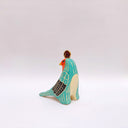 A stylized bird figurine in turquoise, cream, black, red, yellow, and brown, with a gold coin atop its head, photographed on a plain light gray background.
