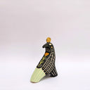 Ceramic sculpture of a black billed magpie, with stylized feather patterning in cream and mint green. On its back is a poppy flower and atop its head is a gold coin.