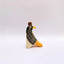 A stylized ceramic red-tailed hawk sculpture, with a black and beige body, yellow beak and tail accents, and a small gold coin on its head, photographed against a plain light gray background.