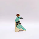 A stylized ceramic bird sculpture in profile, painted with turquoise, black, and cream colors, featuring a small gold circular coin on its head, set against a plain light gray background.