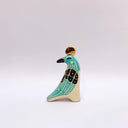 A stylized ceramic bird sculpture in profile, painted with turquoise, black, and cream colors, featuring a small gold circular coin on its head, set against a plain light gray background.