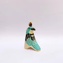 A stylized ceramic bird sculpture in profile, painted with turquoise, black, and cream colors, featuring a small gold circular coin on its head, set against a plain light gray background.