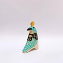 A stylized ceramic bird sculpture in profile, painted with turquoise, black, and cream colors, featuring a small gold circular coin on its head, set against a plain light gray background.