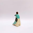 A stylized ceramic bird sculpture in profile, painted with turquoise, black, and cream colors, featuring a small gold circular coin on its head, set against a plain light gray background.