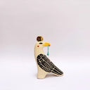 A stylized ceramic bird figurine in cream, black, and yellow, with a small red blush dot on its cheek and a green hanging tassel in its beak, photographed against a plain light gray background.