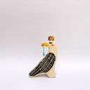 A stylized ceramic bird figurine in cream, black, and yellow, with a small red blush dot on its cheek and a green hanging tassel in its beak, photographed against a plain light gray background.